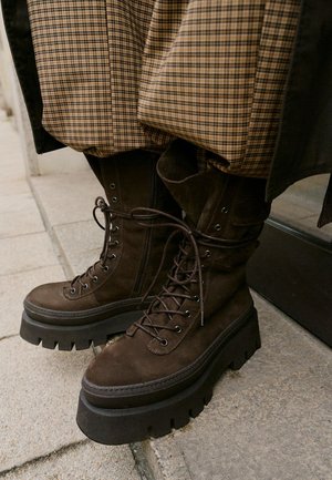 Bruine suède gevechtslaarzen met dikke zwarte zolen, voorzien van een vetersluiting en metalen oogjes, gecombineerd met een pantalon met een geruit patroon.