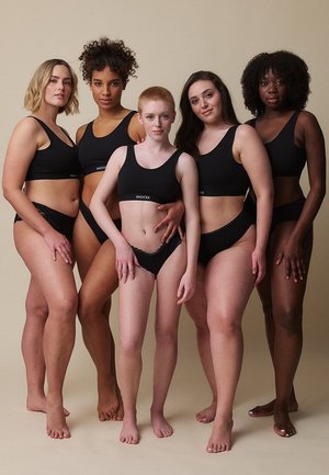 Vijf diverse vrouwen staan op blote voeten, gekleed in zwarte sportbeha's en ondergoed, en poseren vol zelfvertrouwen tegen een neutrale achtergrond.