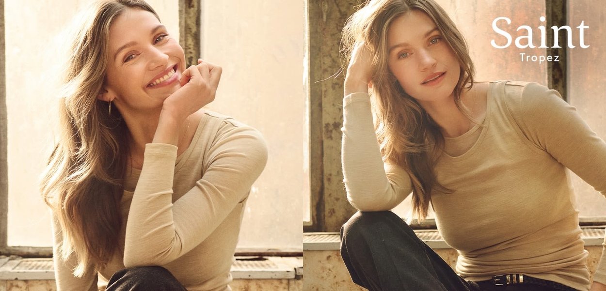 Top beige chiaro a maniche lunghe con scollo rotondo; il modello presenta onde morbide e trucco discreto, seduto accanto a una finestra con luce naturale soffusa.