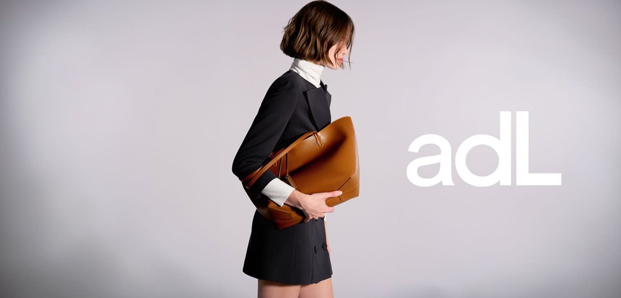 Bruine leren tas met een gestructureerde vorm, voorzien van een gladde textuur en zichtbare stiksels. Model draagt een zwarte outfit en een witte kraag.