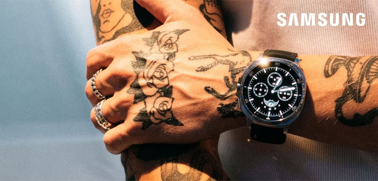 Schwarze Smartwatch an einem tätowierten Handgelenk mit einem runden Zifferblatt, silbernen Akzenten, mehreren Ziffernblättern und einem schwarzen Armband. "SAMSUNG"-Logo in der Ecke.