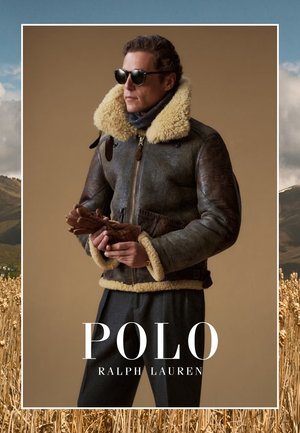 Uomo che indossa una giacca di pelle shearling marrone, occhiali da sole e tiene in mano dei guanti di pelle marroni su uno sfondo beige uniforme.
