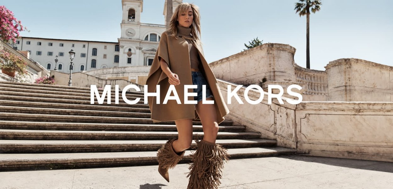 Beige poncho med høy krage, kombinert med denimshorts og frynseboots, foran en historisk trapp med planter og arkitektur i bakgrunnen.