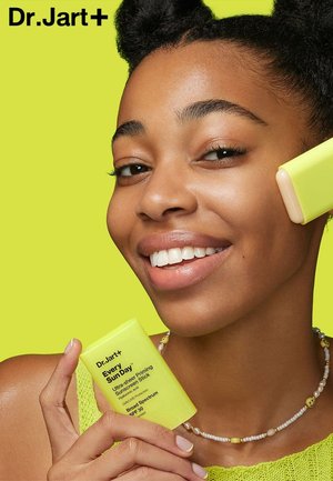 Dr. Jart+ Every Sunday sunscreen stick in bright yellow packaging, held by a person with a pearl necklace, set against a vibrant green background.
