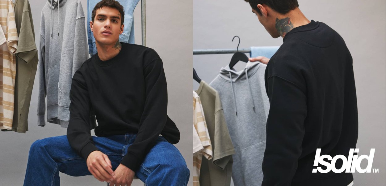 Schwarzer Pullover mit langen Ärmeln, kombiniert mit blauen Jeans. Im Hintergrund hängen ein grauer Kapuzenpullover sowie olivgrüne und gestreifte Hemden an einer Stange.