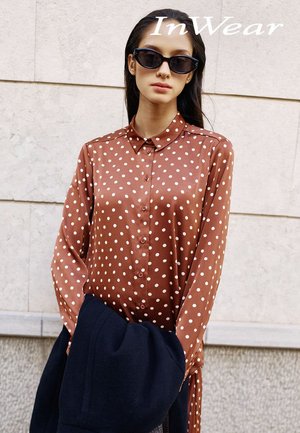 Chemise en satin marron à pois blancs, boutonnée sur le devant, manches longues. Le modèle tient un manteau bleu marine foncé. Le fond est en pierre claire.