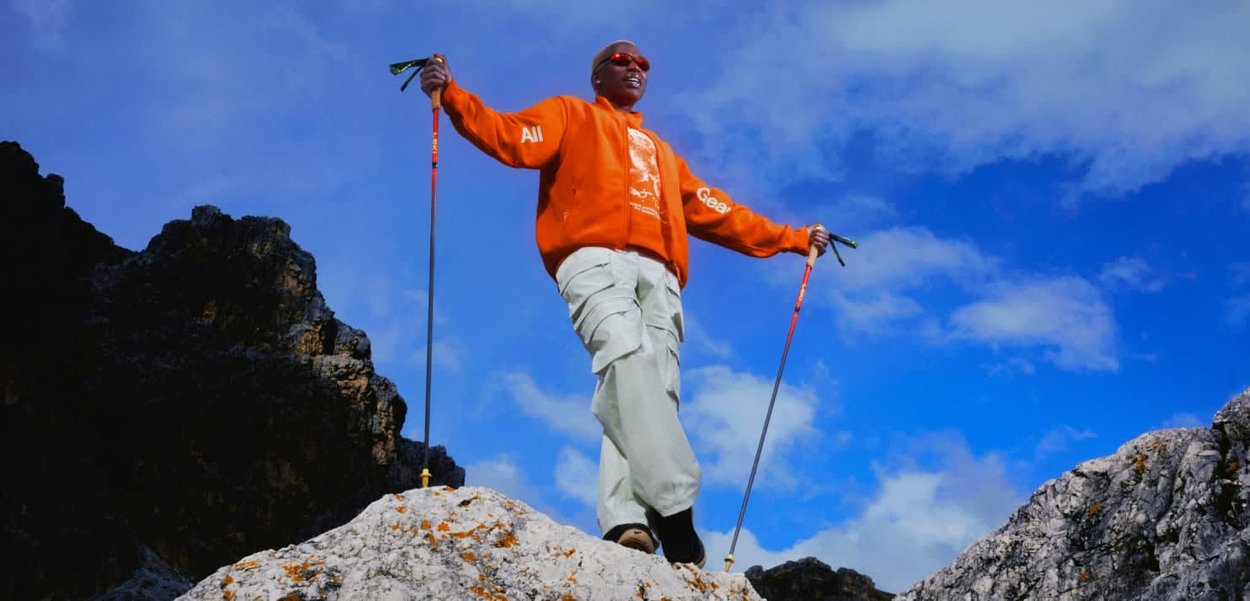 Person mit oranger Jacke und hellen Hosen steht auf felsigem Gelände und hält Trekkingstöcke, unter blauem Himmel mit vereinzelten Wolken.