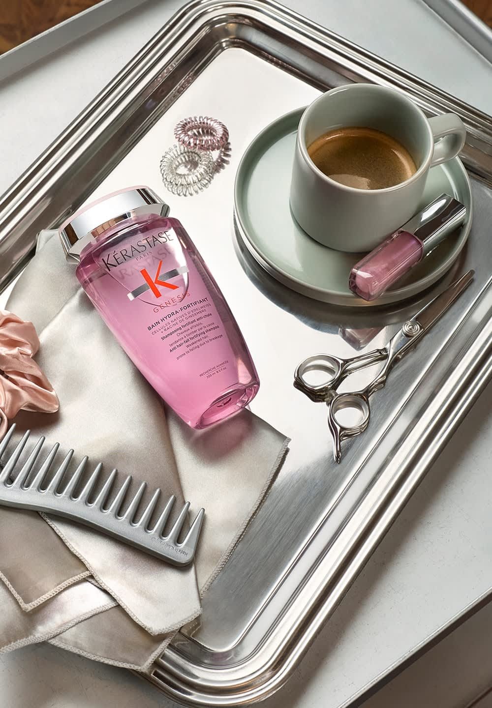 Kérastase pink shampoo bottle, silver comb, shears, pink nail polish, silk cloth, coffee in green cup on a silver tray.