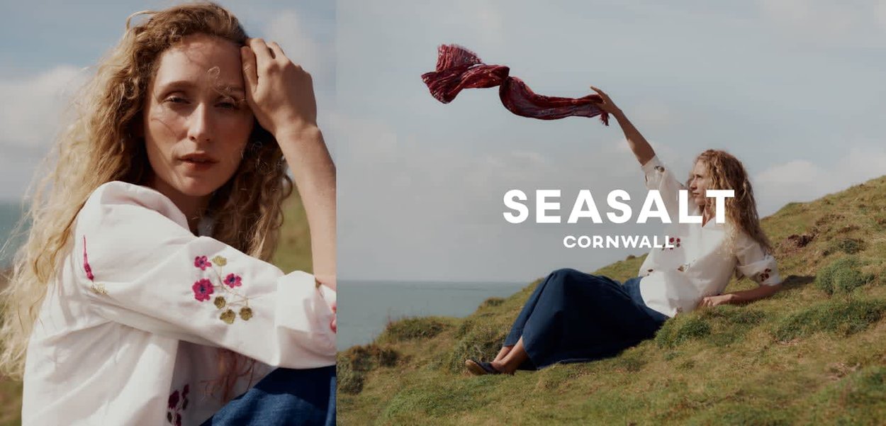 Vrouw met krullend haar draagt een witte bloemenblouse en blauwe jeans, zittend op een grasachtige heuvel aan zee, terwijl ze een rode sjaal in de lucht houdt.