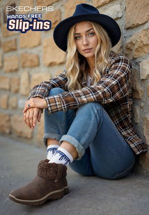 Brown suede ankle boots with a textured cuff, worn with blue jeans and white patterned socks, against a stone wall backdrop.