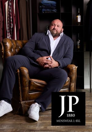 Colbert in marineblauwe stof, witte blouse, zittend in een bruin leren fauteuil. Op de achtergrond zijn kleding en flessen wijn te zien. Logo weergegeven.