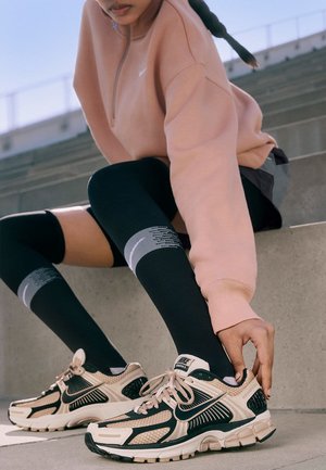 Baskets de sport beiges et noires avec des accents en maille, des lacets et une semelle proéminente, portées avec des chaussettes noires montantes jusqu'au genou avec des rayures grises.