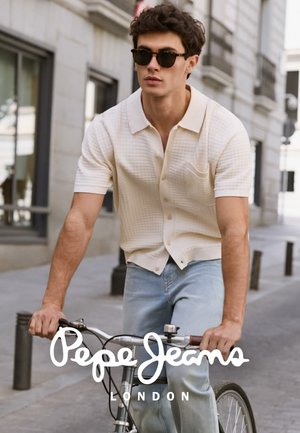 Joven con gafas de sol, camisa blanca de manga corta y vaqueros montando una bicicleta por una calle de la ciudad.
