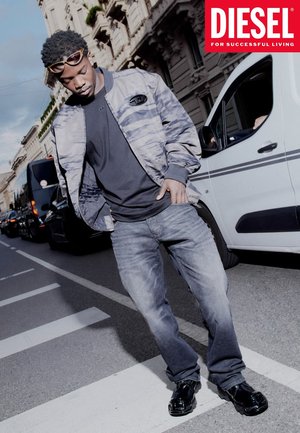 Model trägt eine camouflagemusterte graue Bomberjacke über einem schwarzen T-Shirt, grauen Jeans und schwarzen Schuhen, posiert auf einer Straße.