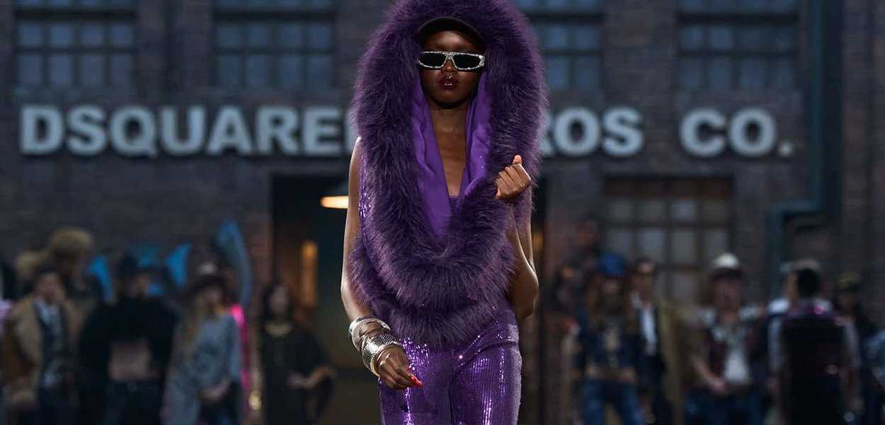 Paarse pailletten outfit met een capuchon met bontkraag. Model draagt een zonnebril, rode nagellak en zilveren armbanden, lopend op een catwalk.