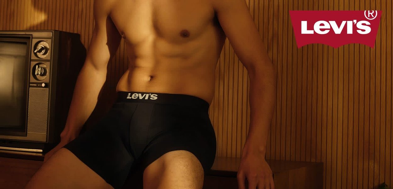Een blote man in zwarte Levi's boxershorts, zittend op houten meubels naast een oude televisie in een kamer met houten paneeltjes.