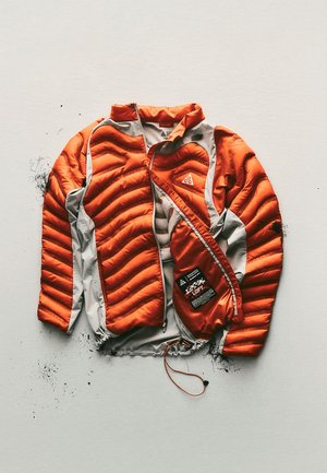 Giacca da esterno isolata arancione e grigia con imbottitura visibile e logo sul petto, distesa su uno sfondo chiaro con un po' di sporco.