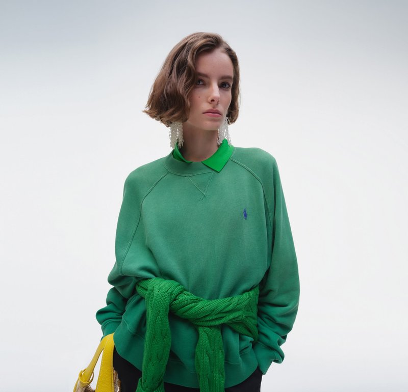 Mujer con cabello corto y castaño lleva sudadera verde sobre camisa verde con cuello y grandes pendientes, sosteniendo un bolso amarillo sobre fondo blanco.