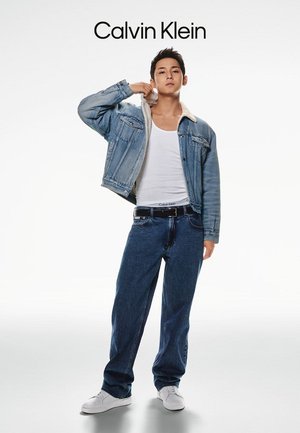 Jeansjacke mit Lammfellkragen, weißem Tanktop, blauen Jeans und weißen Sneakers. Mit Calvin Klein-Logo am Bund.