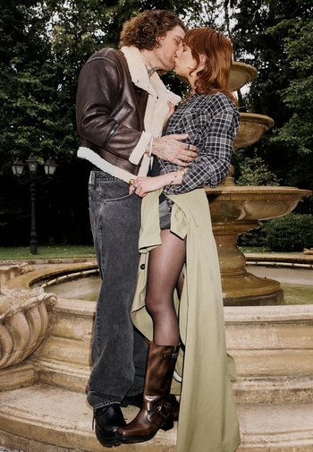 Due persone si baciano vicino a una fontana. L'uomo indossa una giacca foderata in shearling marrone, jeans grigi e stivali neri. La donna ha una camicia a fantasia e un cappotto verde chiaro.