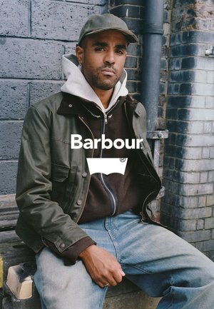 Grüne gewachste Jacke über einem braunen Hoodie, kombiniert mit blauen Jeans. Das Model sitzt an einer Ziegelmauer und zeigt einen lässigen urbanen Stil.