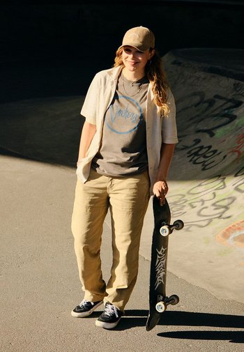 Skateboard tenue par une personne portant un t-shirt gris, une chemise légère à boutons, un pantalon cargo beige et des baskets noires dans un skate park.