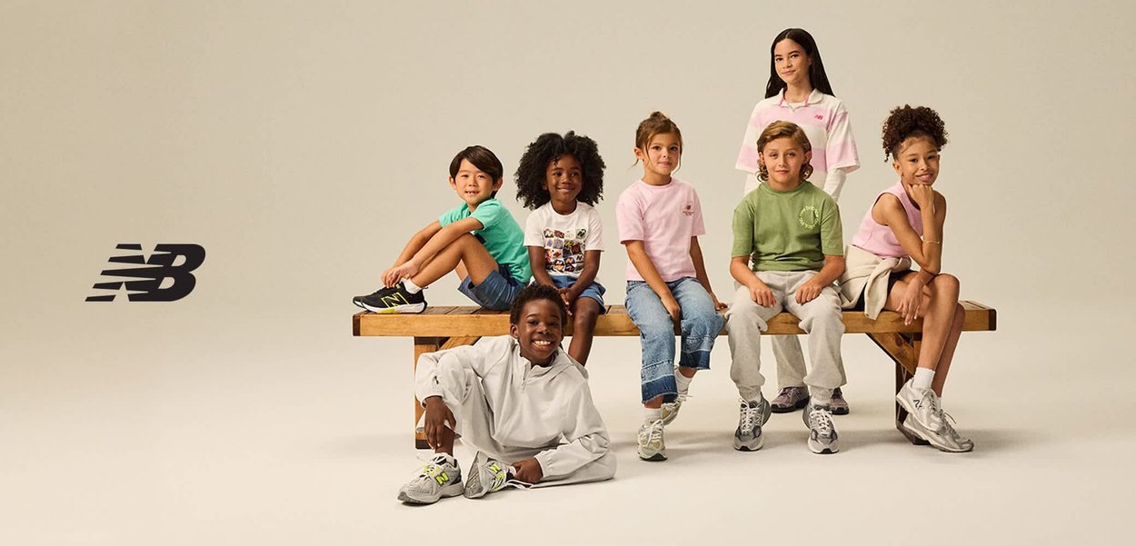 Zeven kinderen in casual New Balance-kleding en sneakers poseren op en rond een houten bank tegen een effen achtergrond.