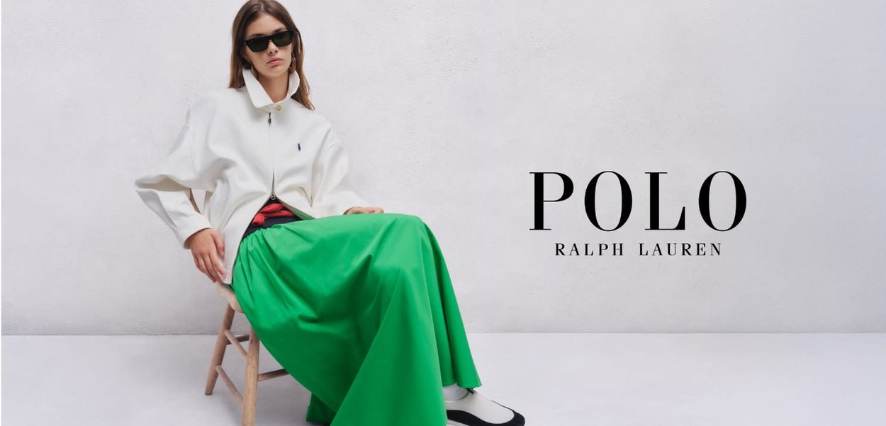 Mujer con gafas de sol lleva una chaqueta blanca y una falda larga verde, sentada en una silla de madera contra un fondo claro y simple.