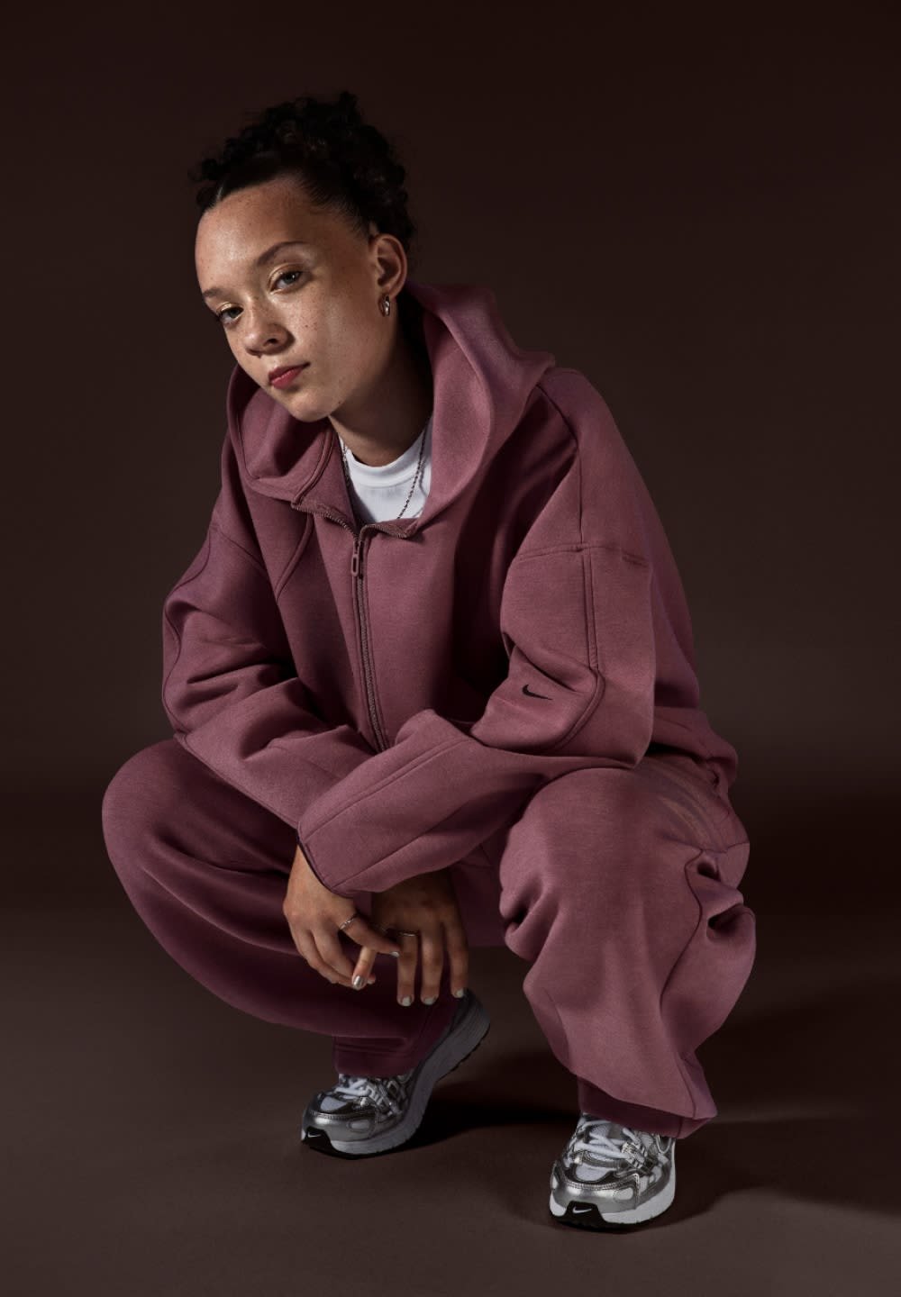 Ung person i knæbøjning iført oversize mauve hættetrøje og joggingbukser med sølvfarvede sneakers foran en mørkebrun baggrund.