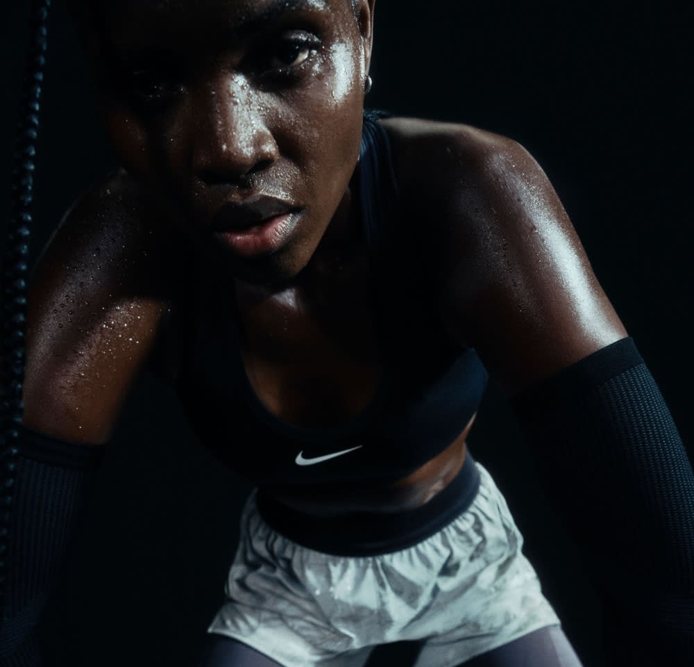 Soutien-gorge de sport noir et shorts gris clair ; la peau brille de transpiration. Gros plan sur un visage concentré avec des traits faciaux bien définis sur fond sombre.