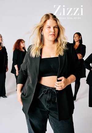 Une femme confiante en crop top noir et blazer se tient devant, avec quatre femmes diverses en tenues noires derrière elle sur un fond blanc.