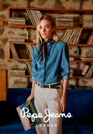 Giovane donna in camicia di jeans e sciarpa che tiene un disco in vinile, in piedi davanti a scaffali di legno pieni di dischi in una stanza accogliente.