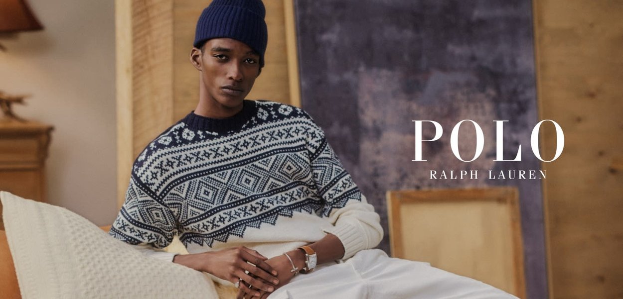 Ung mann iført marineblå genser med mønster og lue slapper av på kremfarget sofa med puter i en koselig innendørs setting.