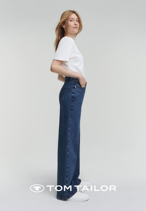 Jeans ampi di denim blu scuro, indossati con una camicia bianca a maniche corte e sneakers bianche, mostrati da una vista laterale.