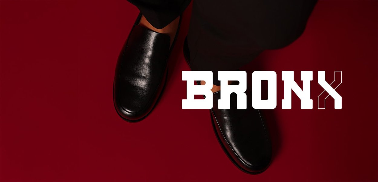 Schwarze Slipper aus Leder mit glatter Textur, präsentiert vor einem roten Hintergrund, mit dem Wort "BRONX" deutlich sichtbar.