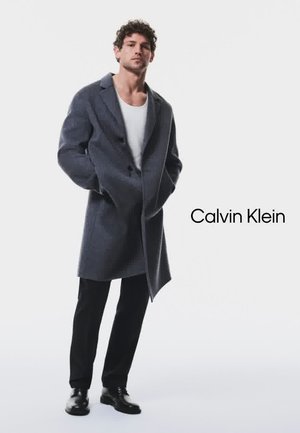 Gray wool blend overcoat with a notched collar, double-breasted buttons, and side pockets, paired with black trousers and shoes.
