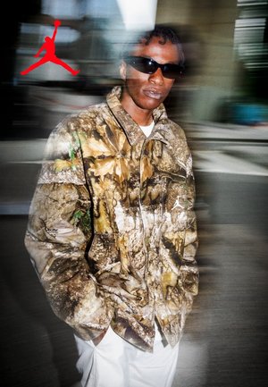 Person wearing sunglasses and a camouflage jacket with hands in pockets, standing in an urban setting with motion blur and a red Jumpman logo.