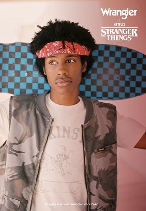 Ein Skateboarder von Wrangler x Stranger Things trägt eine Tarnweste über einem weißen T-Shirt mit Grafik. Er hält ein blau-schwarz kariertes Skateboard.