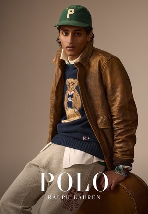 Giacca di pelle marrone, maglione blu con grafica di orso, camicia bianca con colletto, pantaloni del pigiama grigi, cappellino verde con "P", borsa marrone.