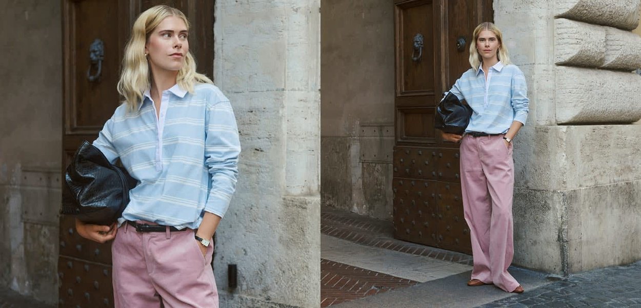 Blonde Frau mit hellblau gestreifter Bluse und pinker Hose, die eine schwarze Tasche hält und neben einer großen Holztür in einem Steingebäude steht.