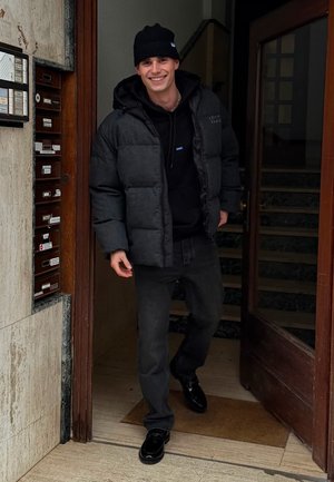 Black puffer jacket with a hood, layered over a black hoodie, paired with black jeans and shiny black shoes. Standing near a wooden door.
