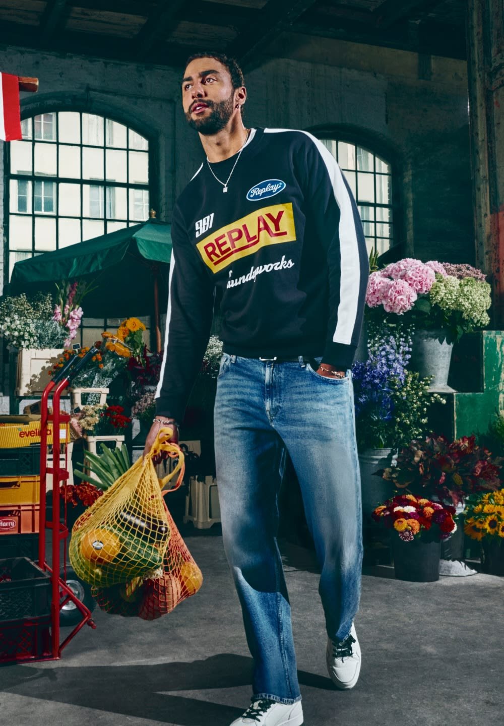 Felpa nera con logo colorato "Replay", strisce bianche, jeans azzurri chiari e sneaker bianche, che tiene in mano una borsa di rete gialla con della frutta.