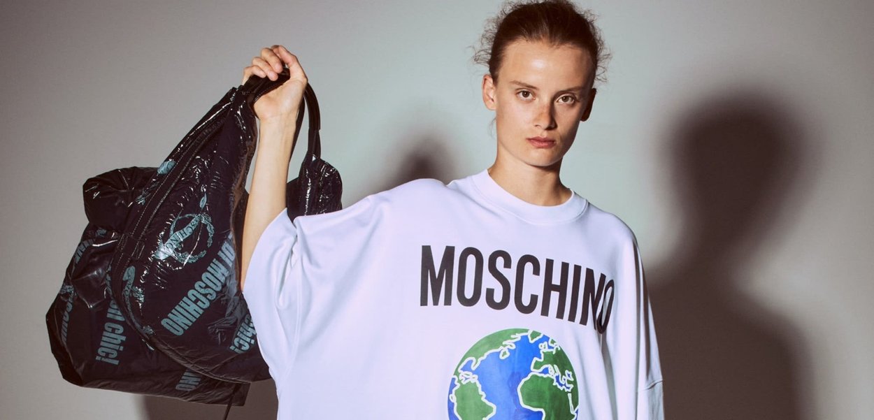 T-shirt bianca oversize con una grafica di un globo blu e verde, testo in grassetto nero "MOSCHINO SOS" e "SAVE OUR SHERE." Il modello tiene in mano una borsa nera.