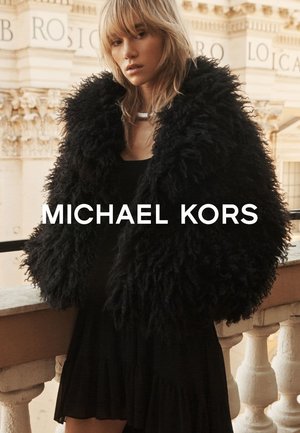 Blonde Frau trägt einen großen schwarzen, strukturierten Pelzmantel und ein schwarzes Kleid, steht auf einem steinernen Balkon mit einem verzierten Gebäude im Hintergrund.