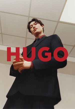 Zwart op maat gemaakt pak met een rood hemd zichtbaar bij de kraag, handen samengevouwen vooraan, en de tekst "HUGO" duidelijk weergegeven in vet rood.