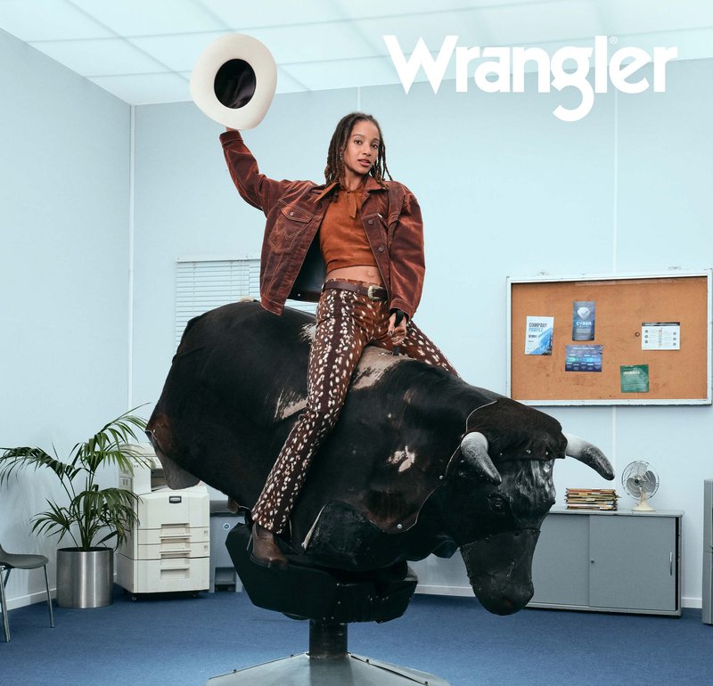 Osoba w wzorzystych spodniach i brązowej kurtce jeździ na mechanicznym byku w pomieszczeniu, trzymając białe kapelusz uniesiony w jednej ręce.