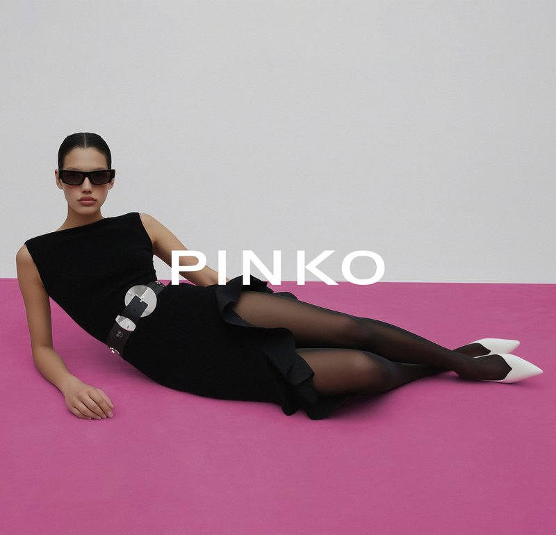 Vrouw liggend op een roze vloer, gekleed in een zwarte jurk met ruches, doorschijnende zwarte panty's, witte puntige schoenen, een oversized riem en zonnebril.