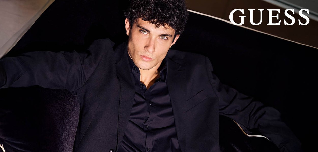 Chaqueta de traje negra sobre una camisa negra, con un cuello clásico. El modelo tiene el cabello corto y rizado y está sentado sobre un tapizado oscuro.