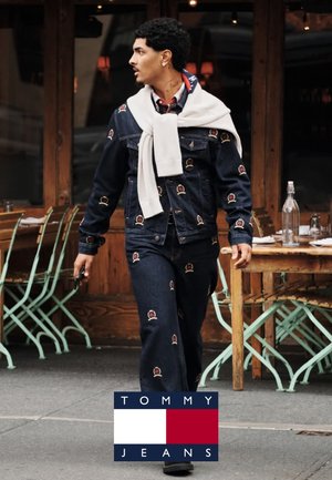 Chaqueta de denim y pantalones con diseños bordados, suéter de punto claro llevado sobre los hombros, caminando por el pavimento cerca de asientos al aire libre.