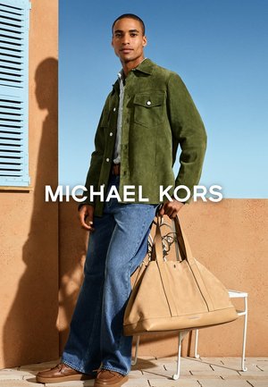 Uomo con giacca di suede verde e jeans in piedi all'aperto con una grande borsa di pelle marrone chiaro contro un muro terracotta e una persiana blu.